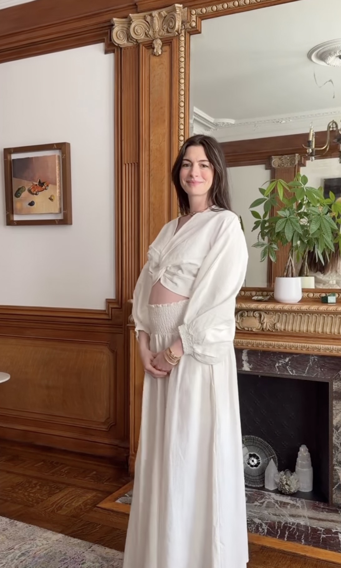 Pregnant woman in white crop top and maxi skirt standing in a wood-paneled room with a ornate gold fireplace and large mirror.