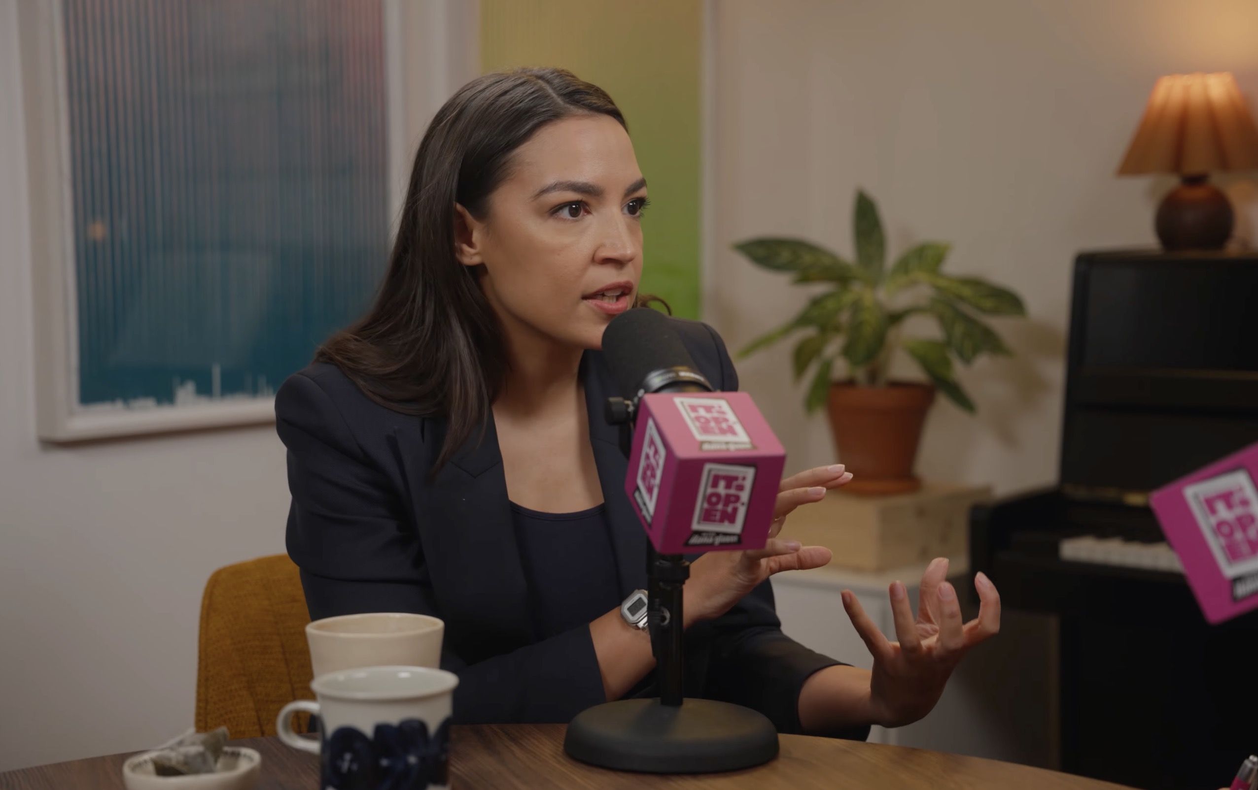 Woman in a navy blazer speaking into a pink branded podcast microphone at a table with coffee cups, gesturing with one hand.