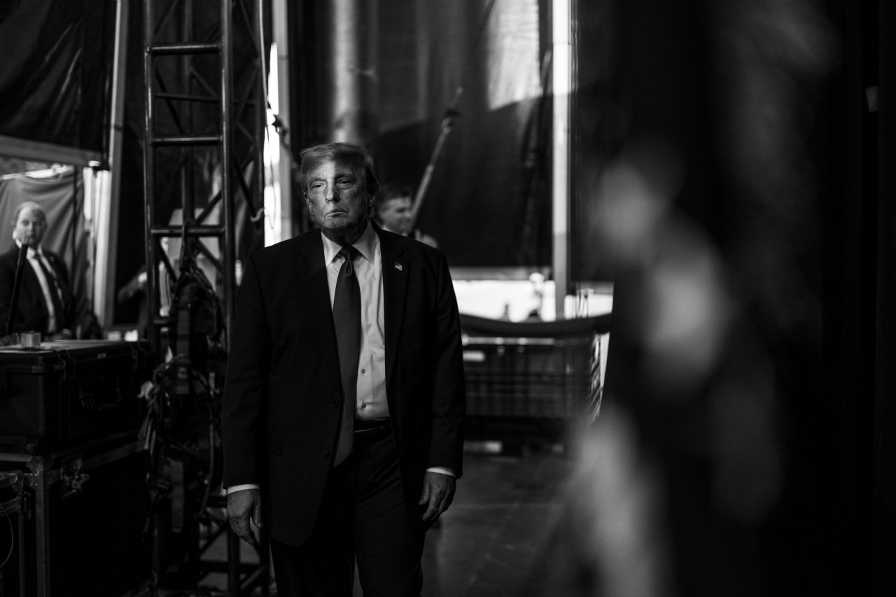 Black and white photo of a man in a dark suit and tie standing backstage amid stage scaffolding and draping, flanked by aides in the background.