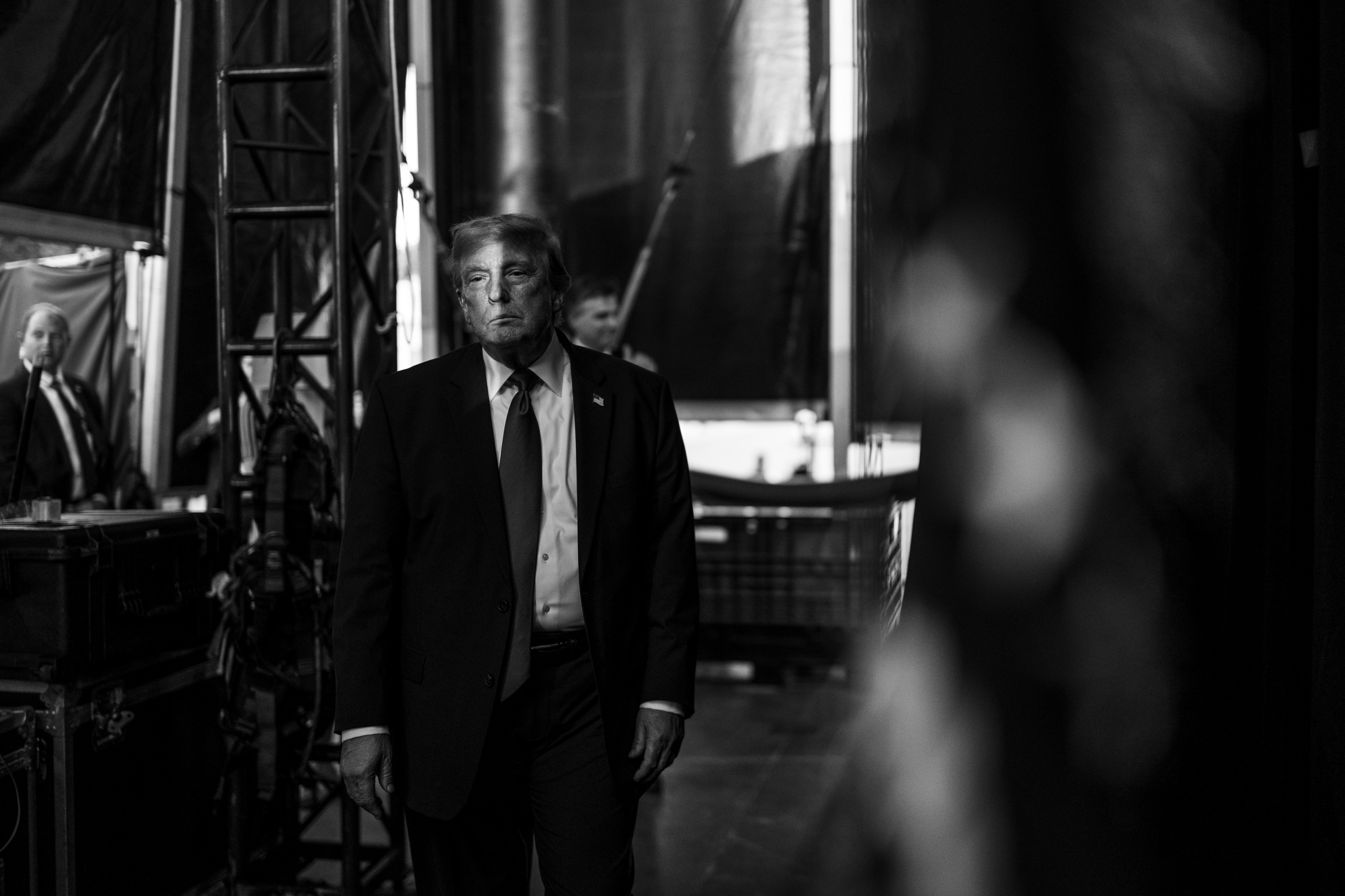 Black and white photo of a man in a dark suit and tie standing backstage amid stage scaffolding and draping, flanked by aides in the background.