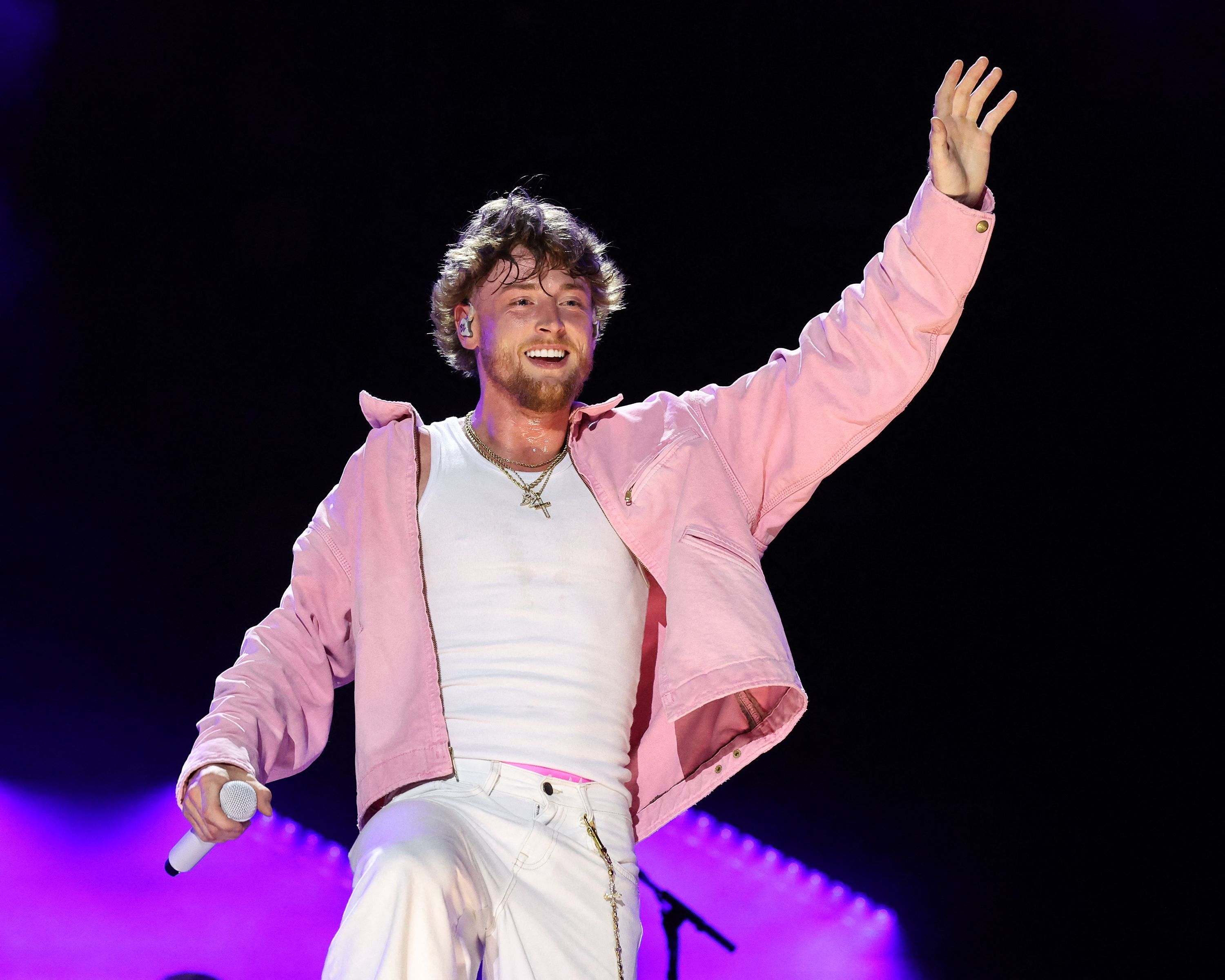 Male performer in a pink jacket and white shirt holding a microphone and waving to the crowd on a darkened stage.