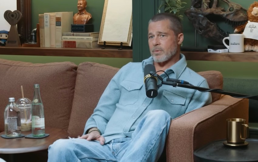 A man with short gray hair and a beard sits on a brown couch wearing a light blue shirt, speaking into a podcast microphone.
