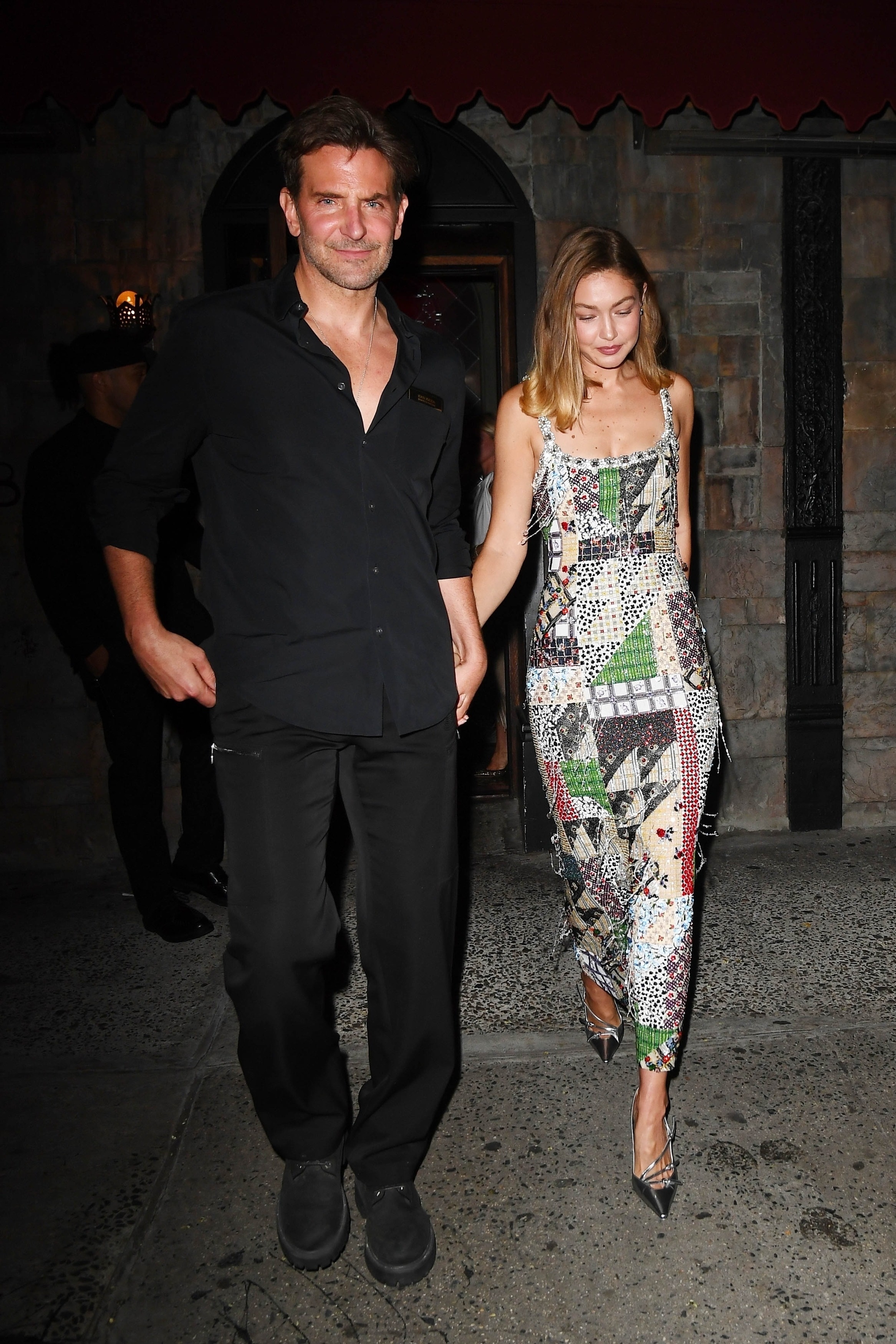 Click to open the image gallery A man in all-black and a woman in a colorful beaded midi dress and silver heels walk hand in hand out of a stone-facade building at night.