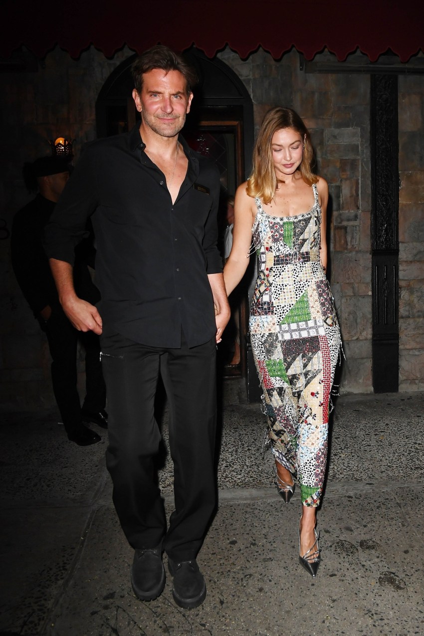 A man in all-black and a woman in a colorful beaded midi dress and silver heels walk hand in hand out of a stone-facade building at night.