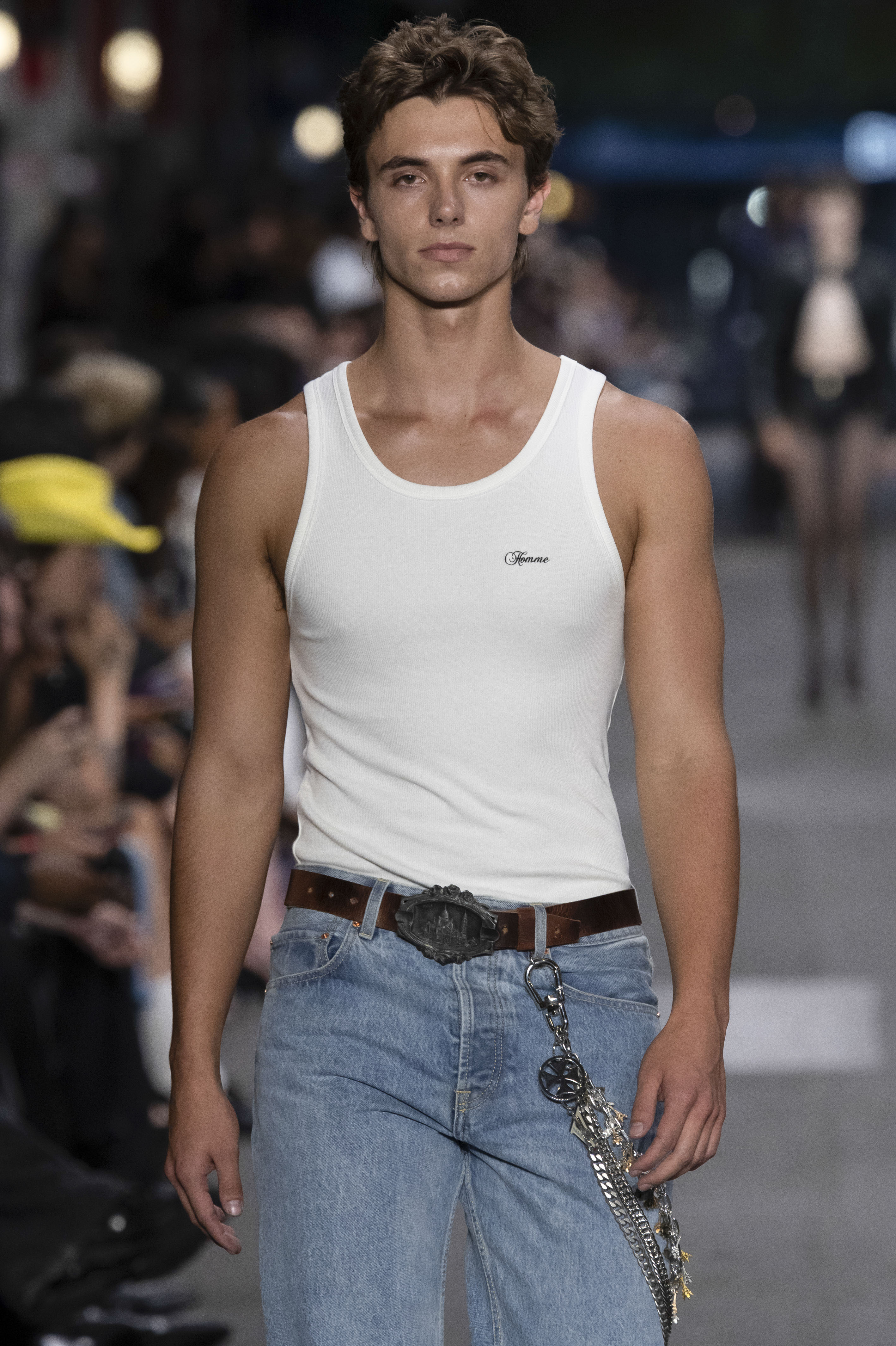 Male model Jayden Federline walks the Vetements runway in a white tank top, light-wash jeans, brown belt with large buckle, and a chain accessory.