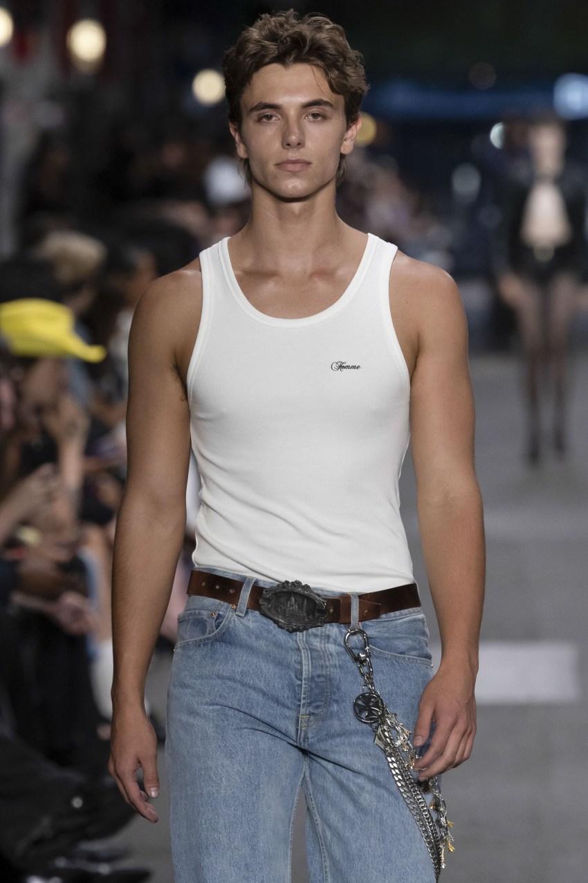 Male model Jayden Federline walks the Vetements runway in a white tank top, light-wash jeans, brown belt with large buckle, and a chain accessory.