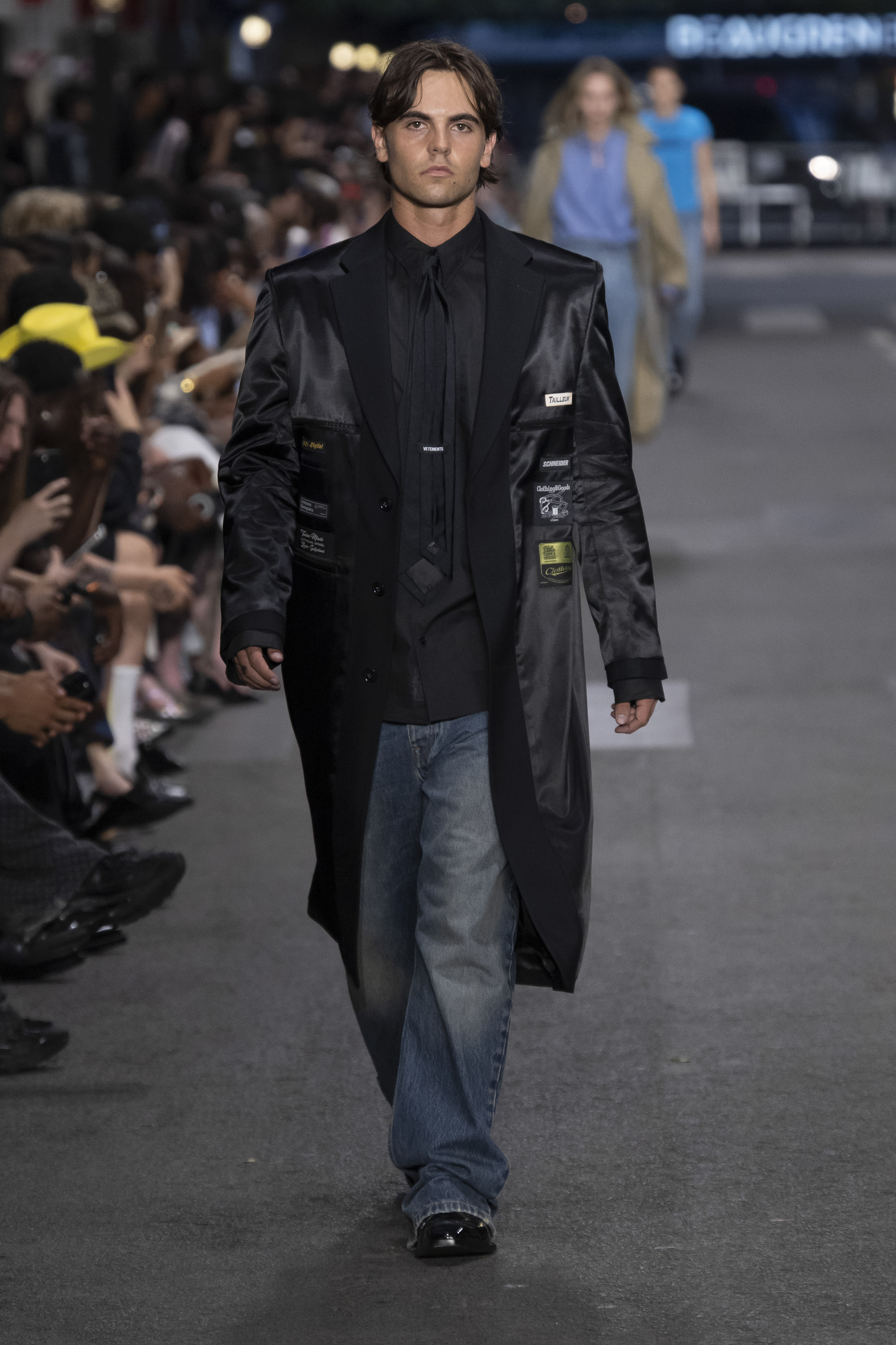 Male model Sean Preston Federline walks the Vetements runway in a long black satin coat with patches, black shirt, and wide-leg jeans.