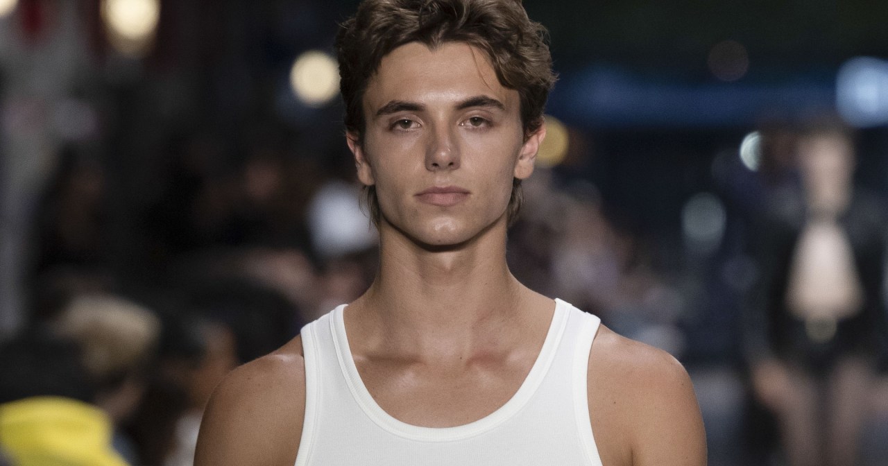 Male model Jayden Federline on a runway in a white tank top, light-wash jeans, brown Western belt, and a chain accessory hanging from his waistband.