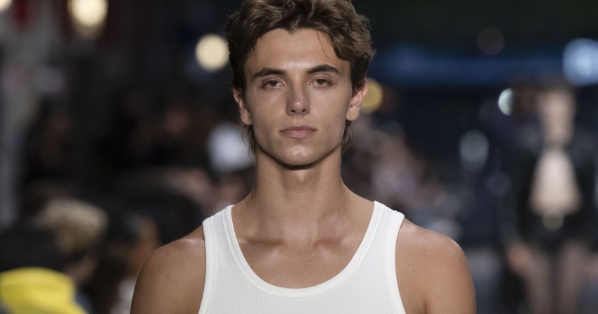 Male model Jayden Federline on a runway in a white tank top, light-wash jeans, brown Western belt, and a chain accessory hanging from his waistband.