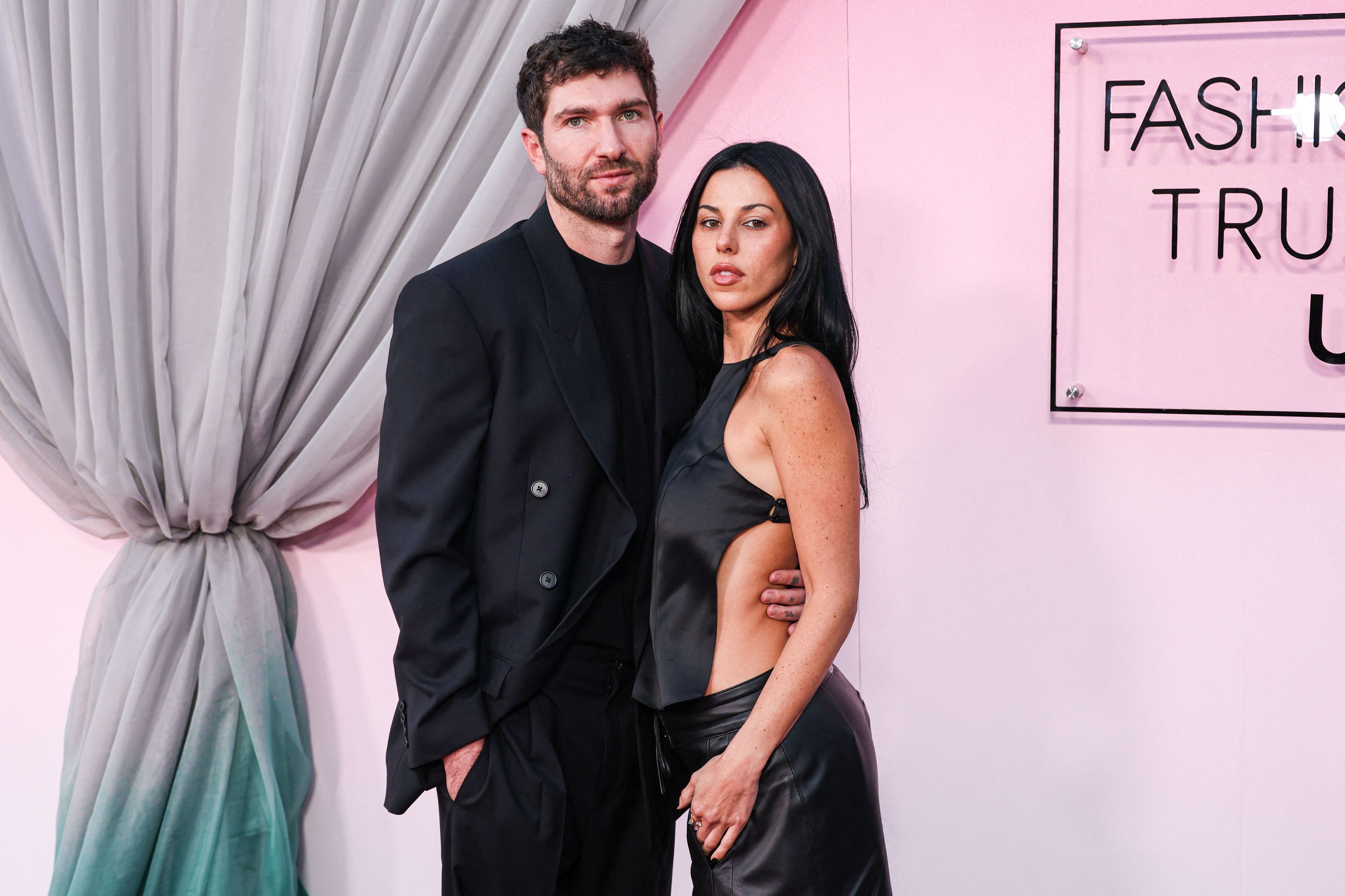 Click to open the image gallery A man in an all-black suit and a dark-haired woman in a black cutout halter top and leather pants posing at a Fashion Trust U.S. Awards event.