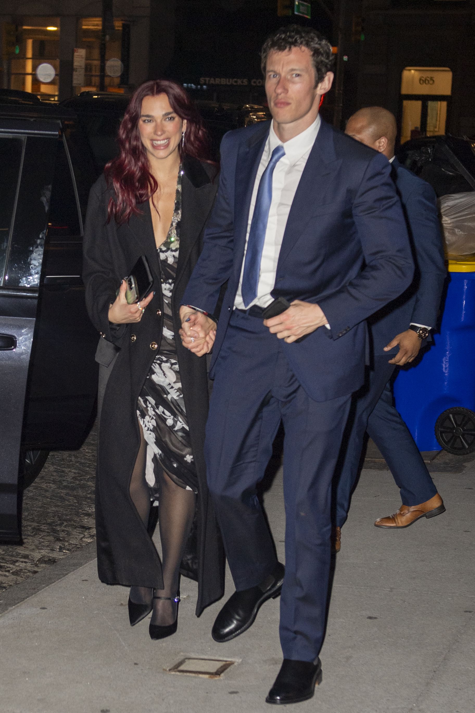 Click to open the image gallery A red-haired woman in a floral dress and black coat walks hand-in-hand with a man in a navy suit on a New York City sidewalk at night.