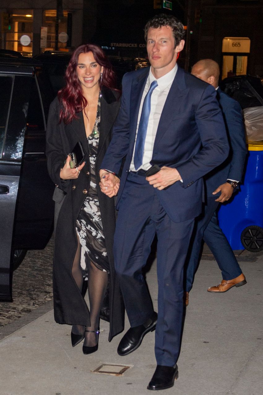 A red-haired woman in a floral dress and black coat walks hand-in-hand with a man in a navy suit on a New York City sidewalk at night.