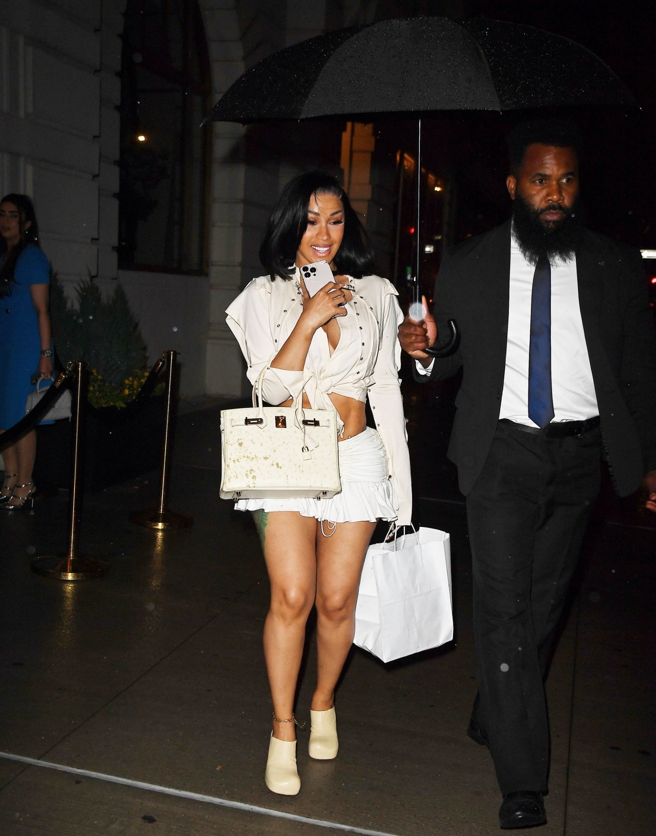 Cardi B in a white cutout top, ruched mini shorts, and cream platform ankle boots holding a white Birkin bag, walking outside at night under a black umbrella held by a suited man.