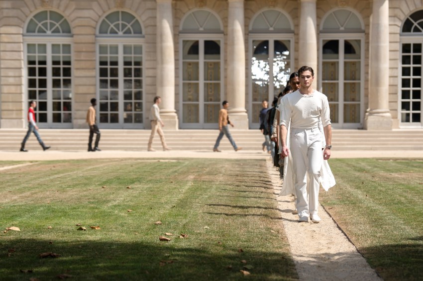 A male model in an all-white outfit leads a line of models walking along a path in front of a neoclassical building during an outdoor fashion show.