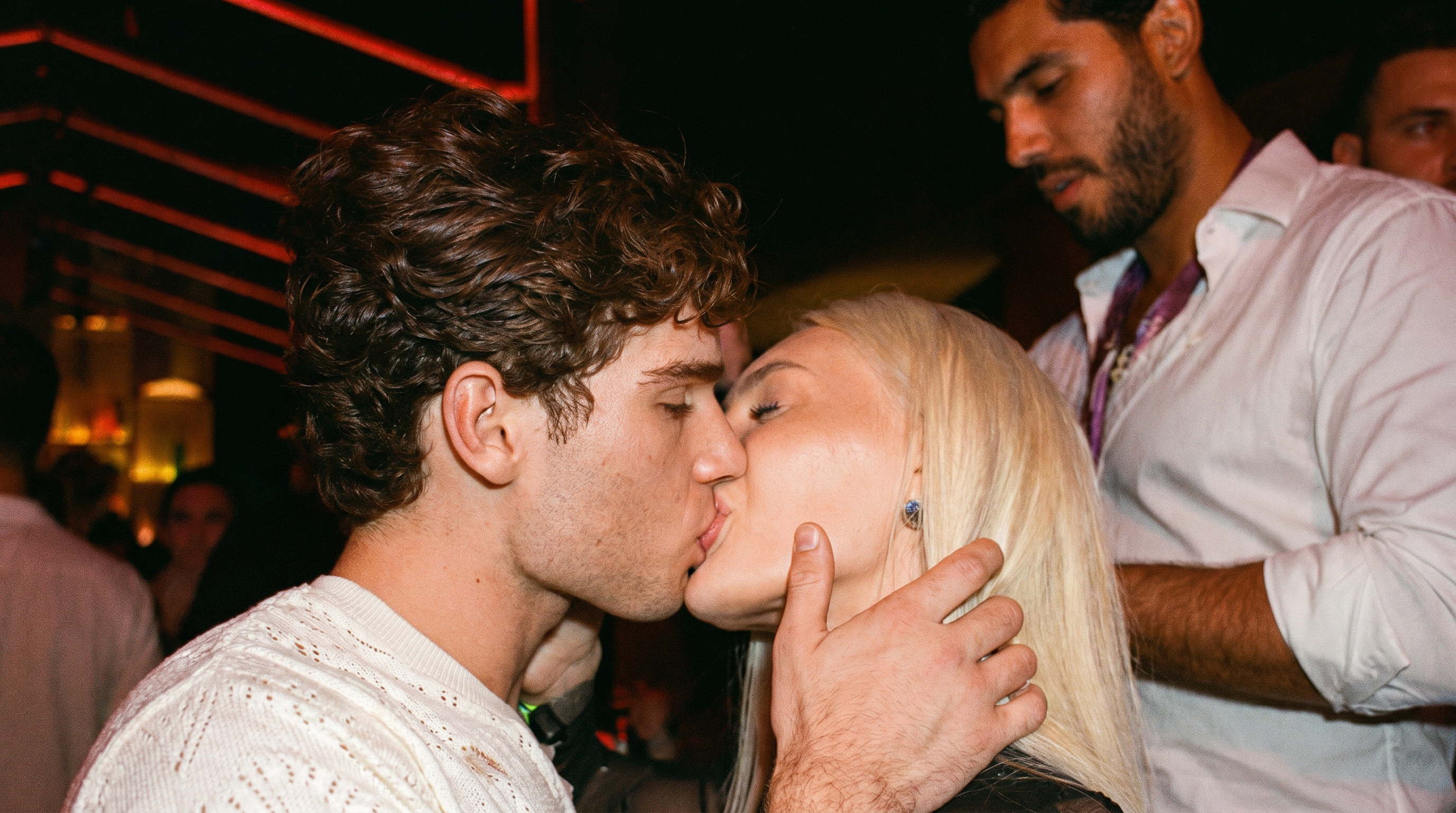 A young man with curly brown hair and a woman with platinum blonde hair kissing at a nightclub.