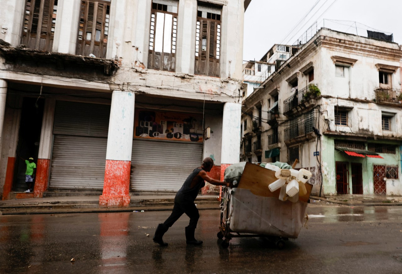 Man pushing a heavily loaded wheelbarrow with plastic containers through wet streets in Havana, Cuba, with deteriorating colonial buildings in the background.