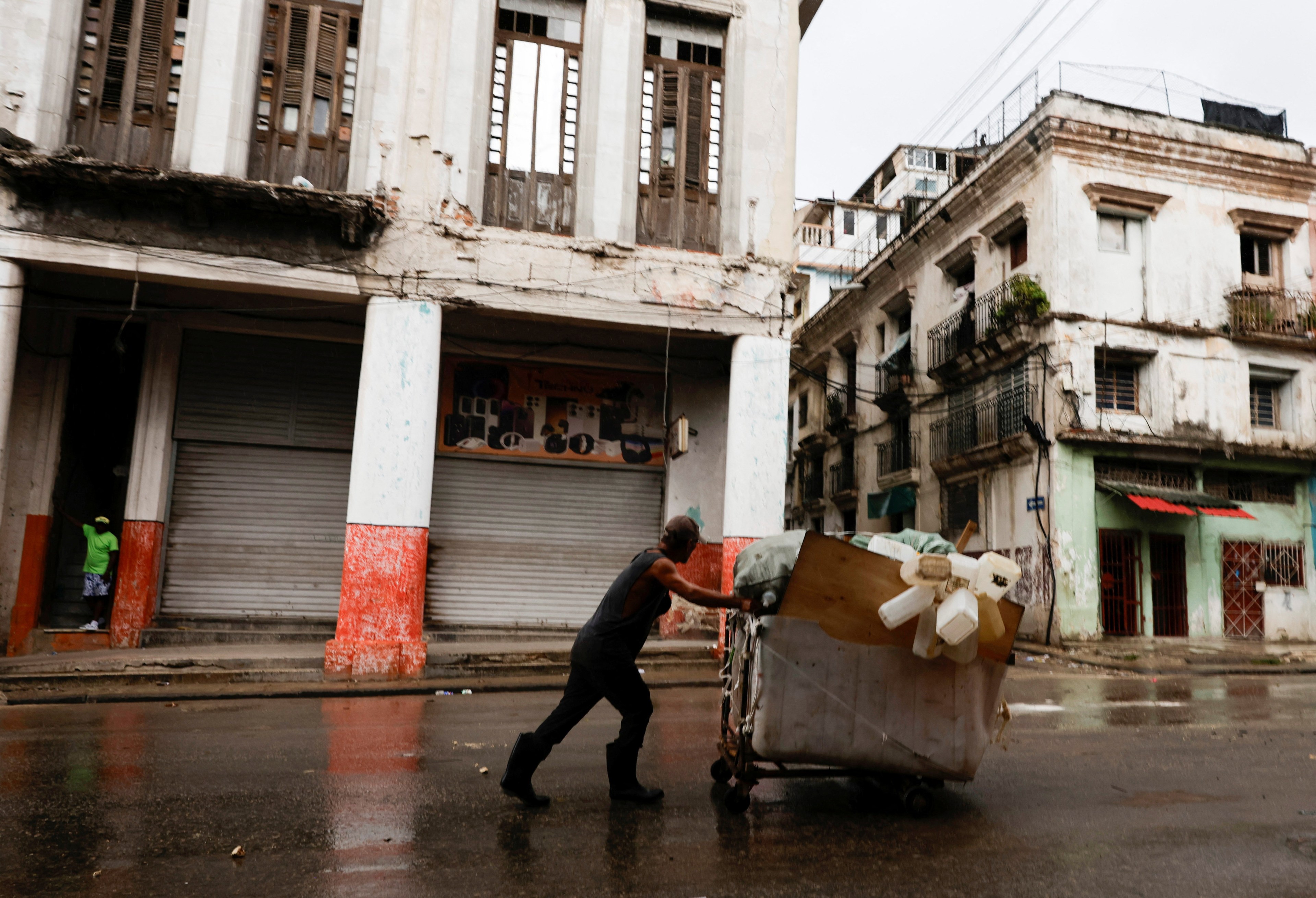 Man pushing a heavily loaded wheelbarrow with plastic containers through wet streets in Havana, Cuba, with deteriorating colonial buildings in the background.