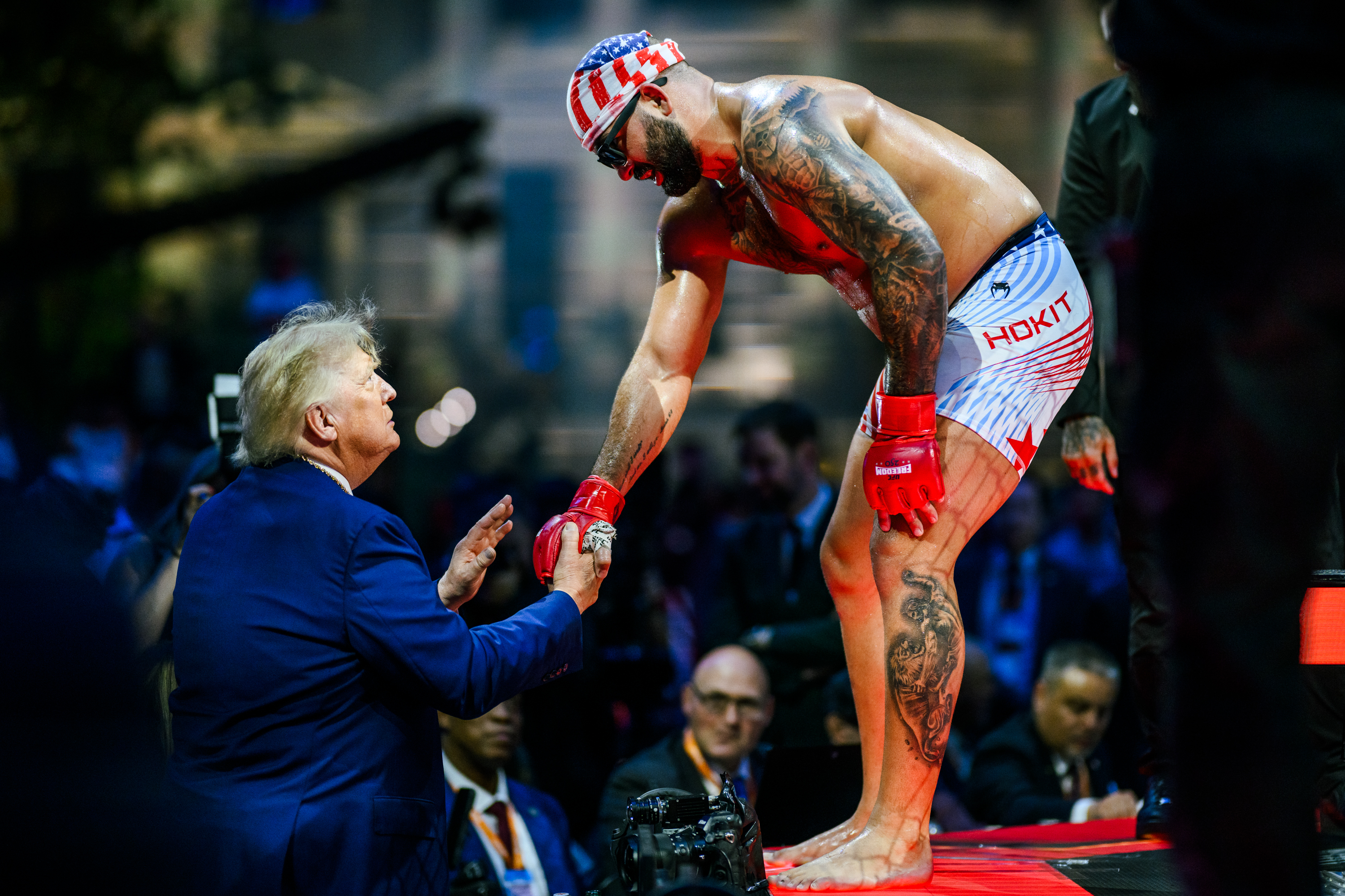 Click to open the image gallery Tattooed MMA fighter in American flag shorts and cap bends down to shake hands with a man in a blue suit at ringside.