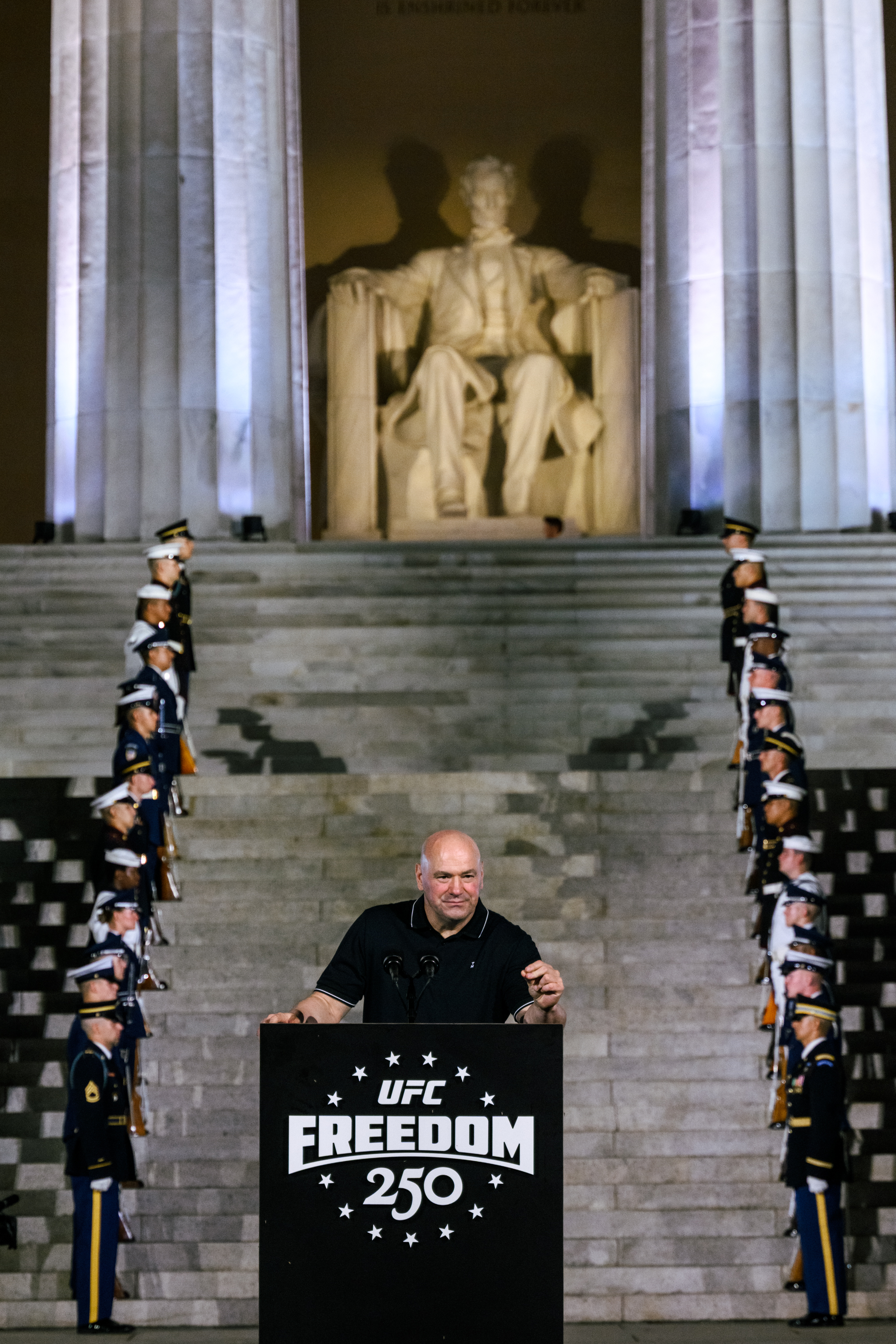 Click to open the image gallery Bald man in black polo speaks at a UFC Freedom 250 podium on the steps of the Lincoln Memorial, with military honor guards lining both sides.