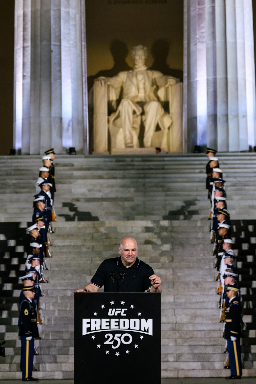 Bald man in black polo speaks at a UFC Freedom 250 podium on the steps of the Lincoln Memorial, with military honor guards lining both sides.