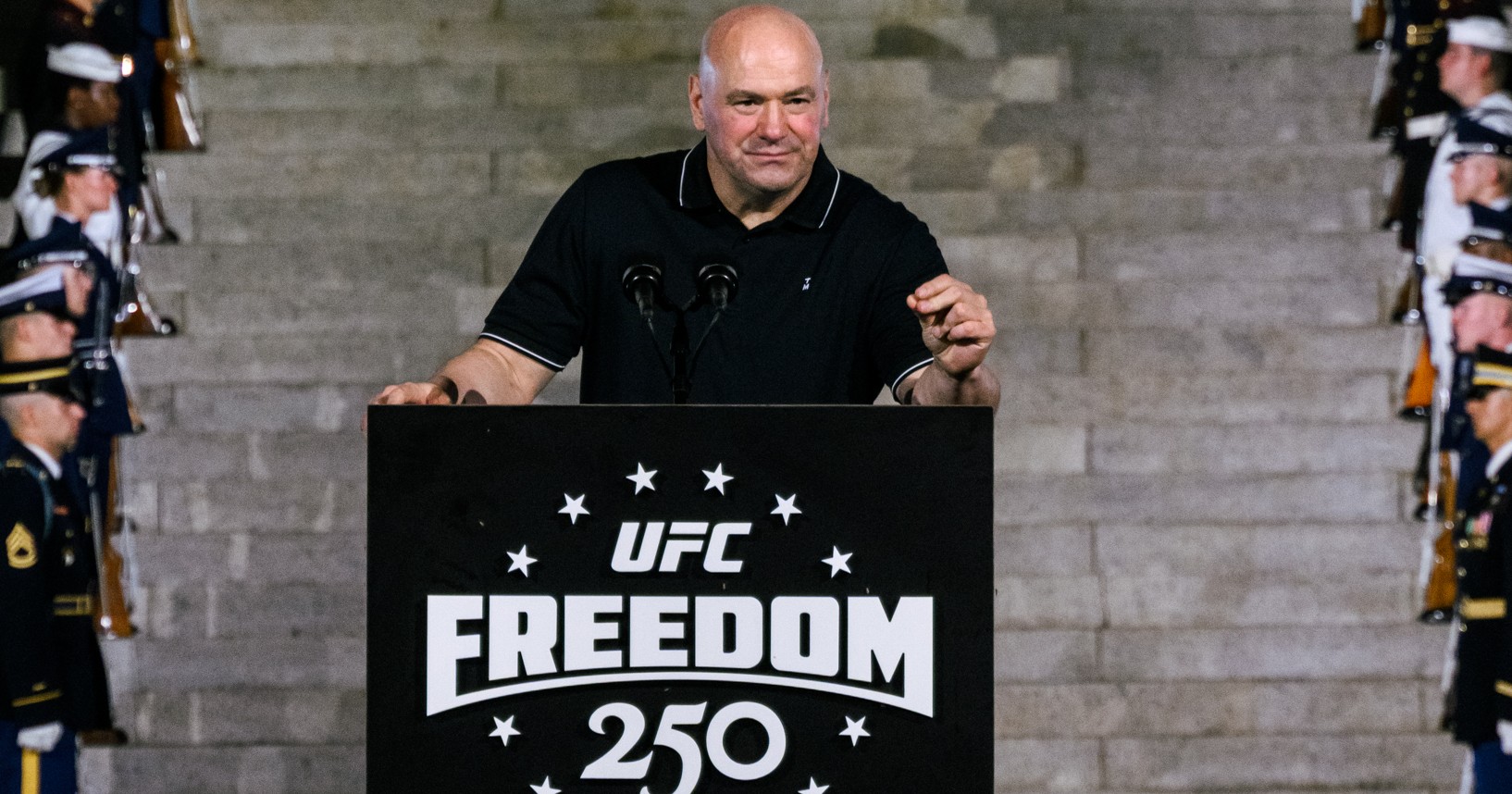 A bald man speaks at a UFC Freedom 250 podium on the steps of the Lincoln Memorial at night, flanked by rows of military honor guards in dress uniforms.
