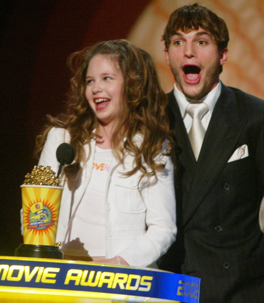 A young curly-haired girl and a man in a black suit with a white tie stand at the MTV Movie Awards podium, both with mouths open in excitement.