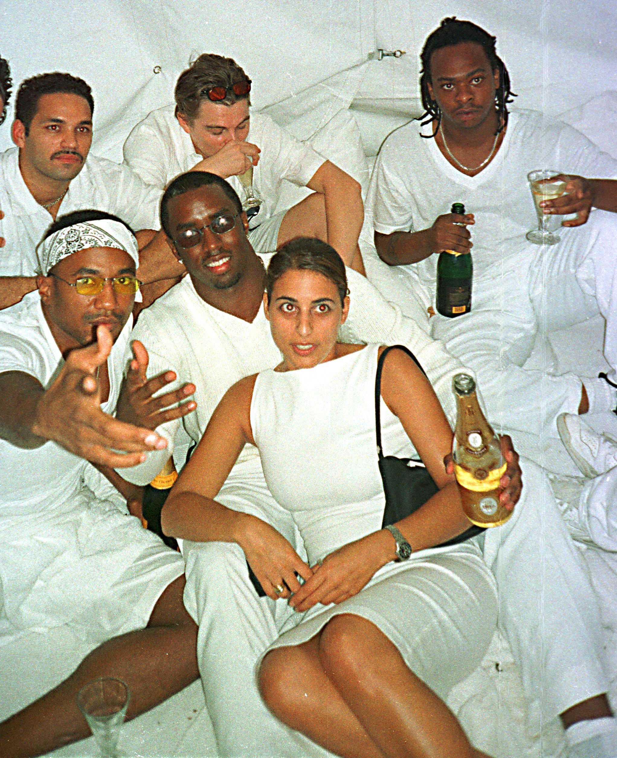 Click to open the image gallery Group of people in all-white outfits lounging together at a party, holding champagne bottles and glasses.