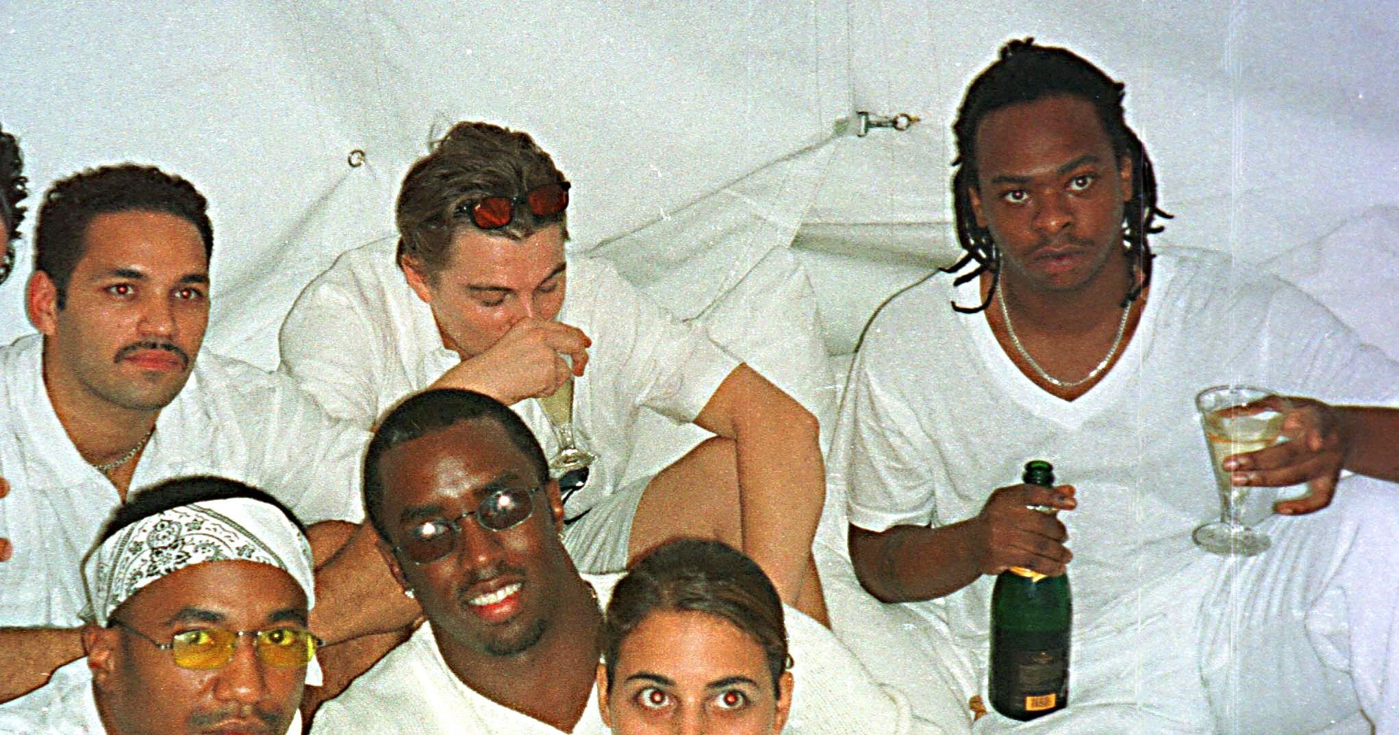 Group of people dressed in white at a party, holding champagne bottles, seated together in a white-draped space.