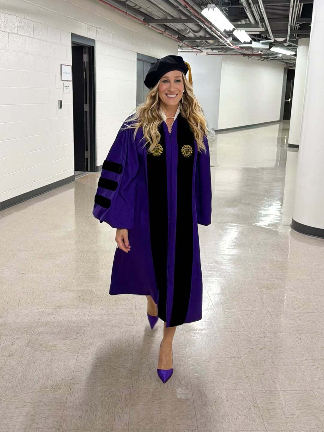 Woman in purple doctoral robes and black tam cap walking confidently down an indoor corridor in matching purple heels.