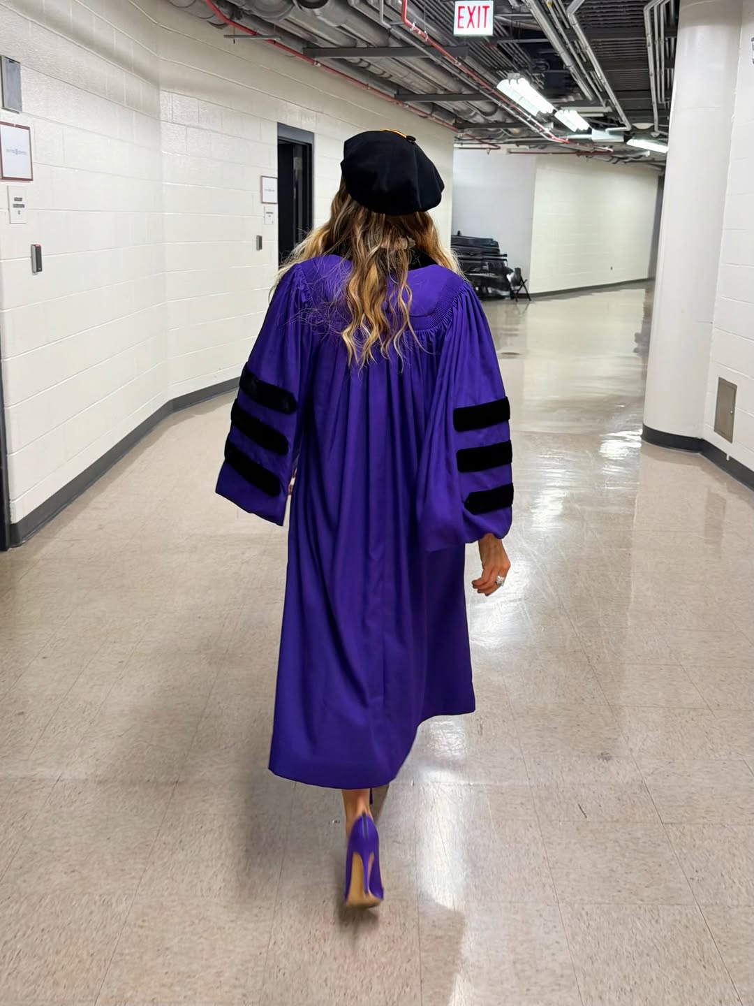 Click to open the image gallery Back view of a woman in a purple doctoral gown with black velvet sleeve stripes and a black tam cap walking down a corridor.