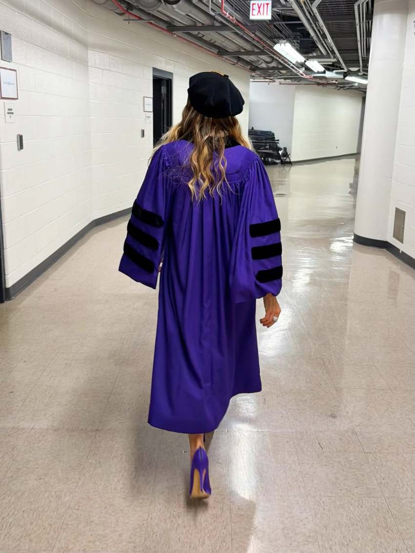 Back view of a woman in a purple doctoral gown with black velvet sleeve stripes and a black tam cap walking down a corridor.