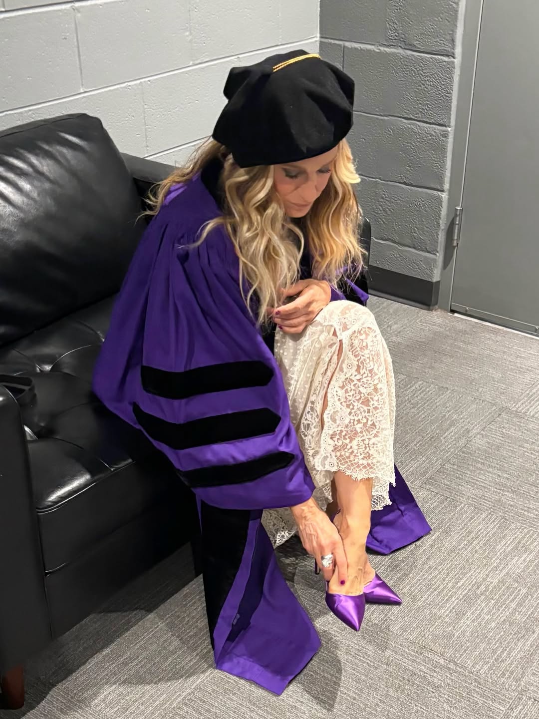 Click to open the image gallery Woman in purple doctoral robes and black tam cap sitting on a leather couch backstage, adjusting a purple satin high heel.