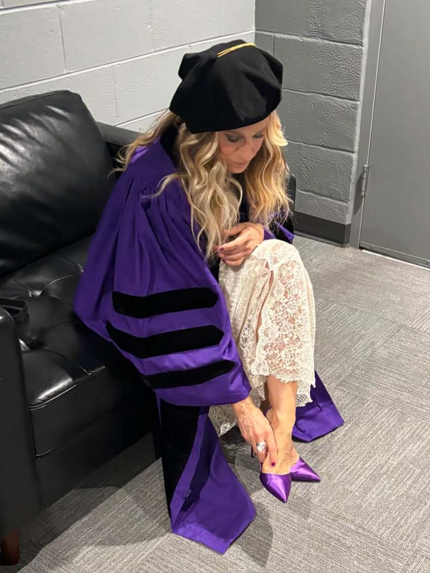 Woman in purple doctoral robes and black tam cap sitting on a leather couch backstage, adjusting a purple satin high heel.