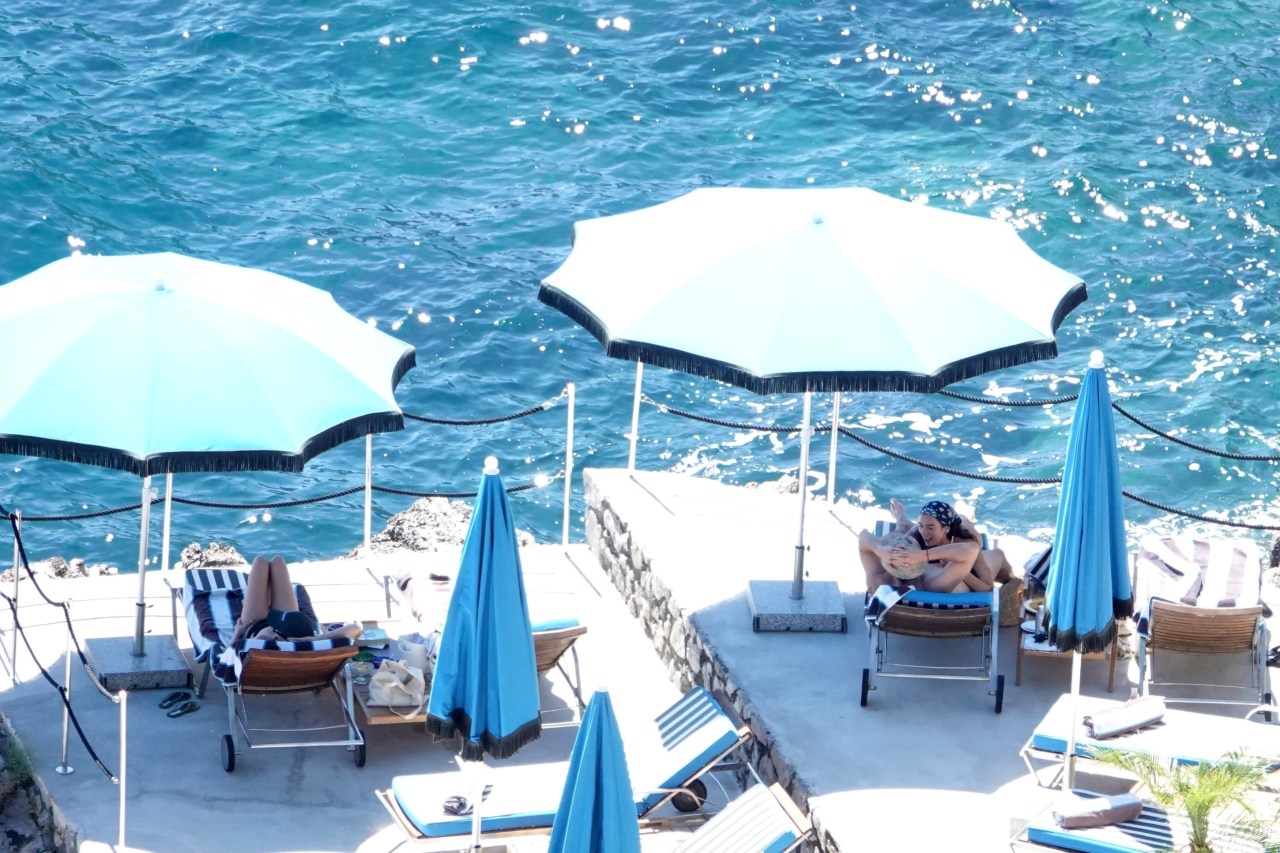 Aerial view of two people embracing on a sun lounger on a stone terrace surrounded by blue fringed parasols, with turquoise sea behind them.