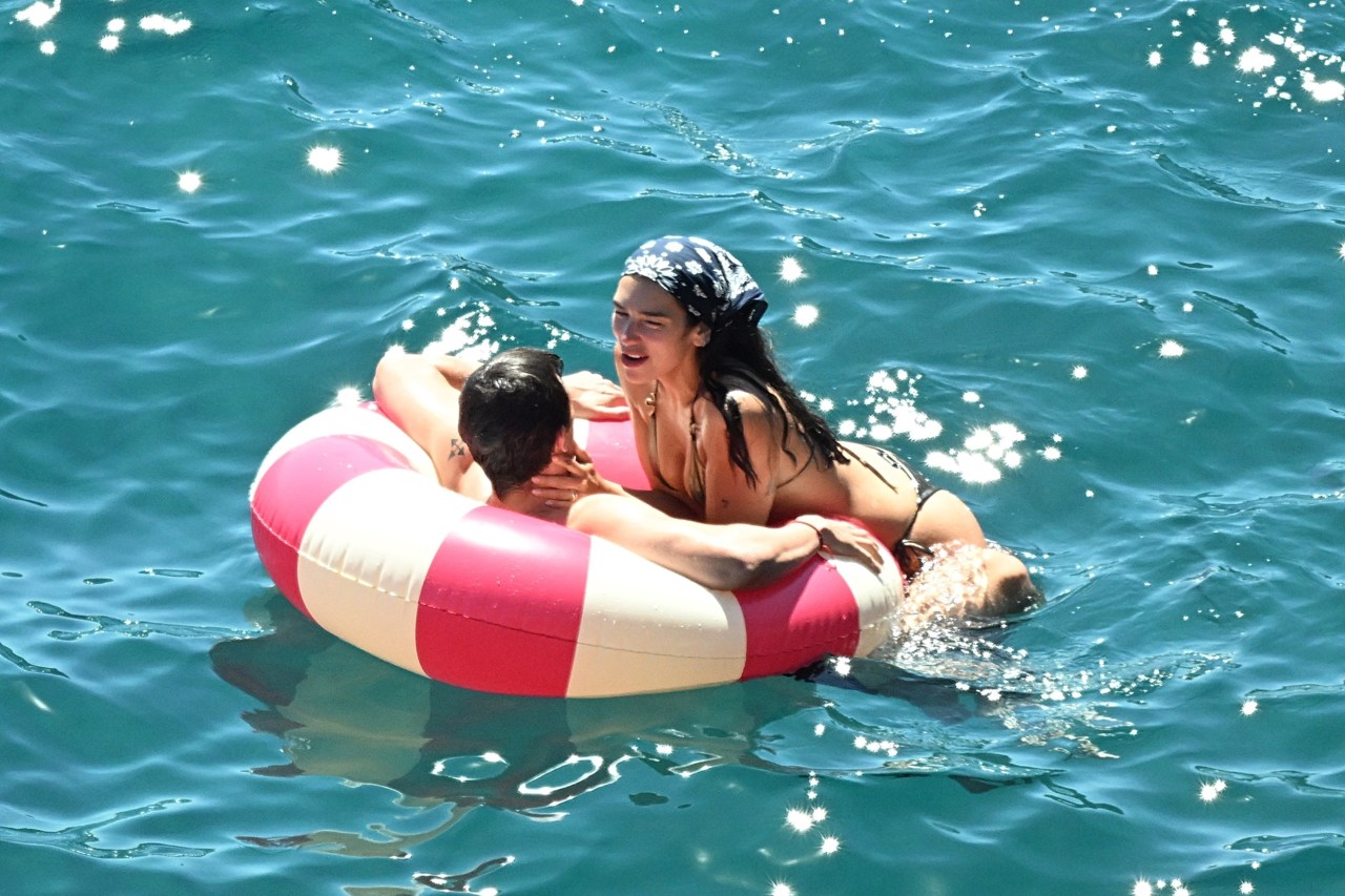 A woman in a dark bikini and head scarf and a man sharing a pink and white inflatable ring in turquoise water, smiling at each other.