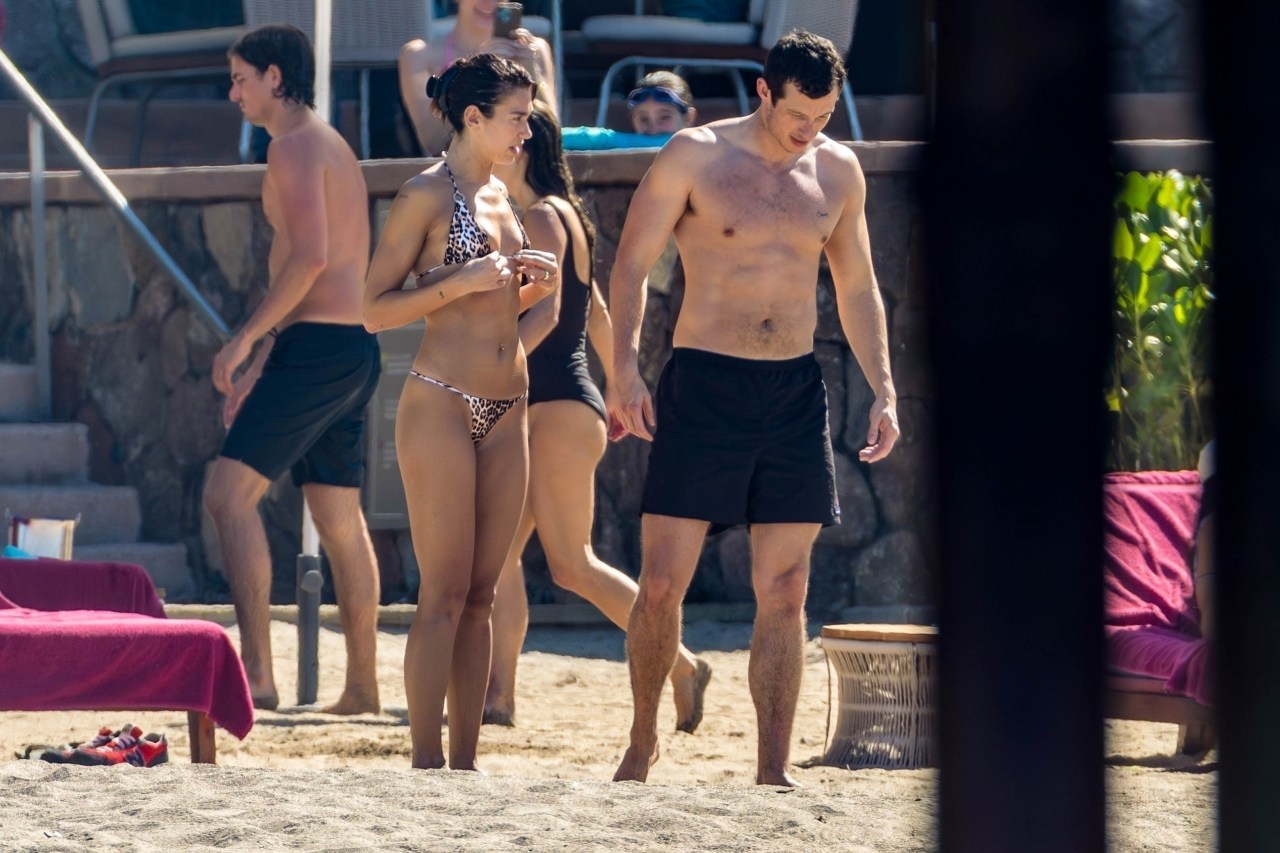 Dua Lipa in a leopard-print bikini walking on a beach alongside a shirtless Callum Turner in black swim shorts, with other beachgoers in the background.