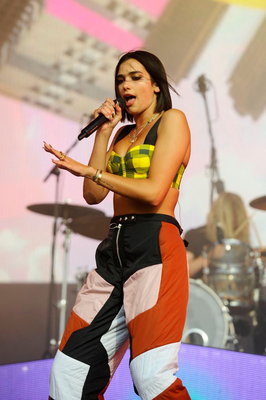 Female singer performing on stage, holding a microphone, wearing a yellow plaid crop top and colorblock pants, with a drum kit visible in the background.