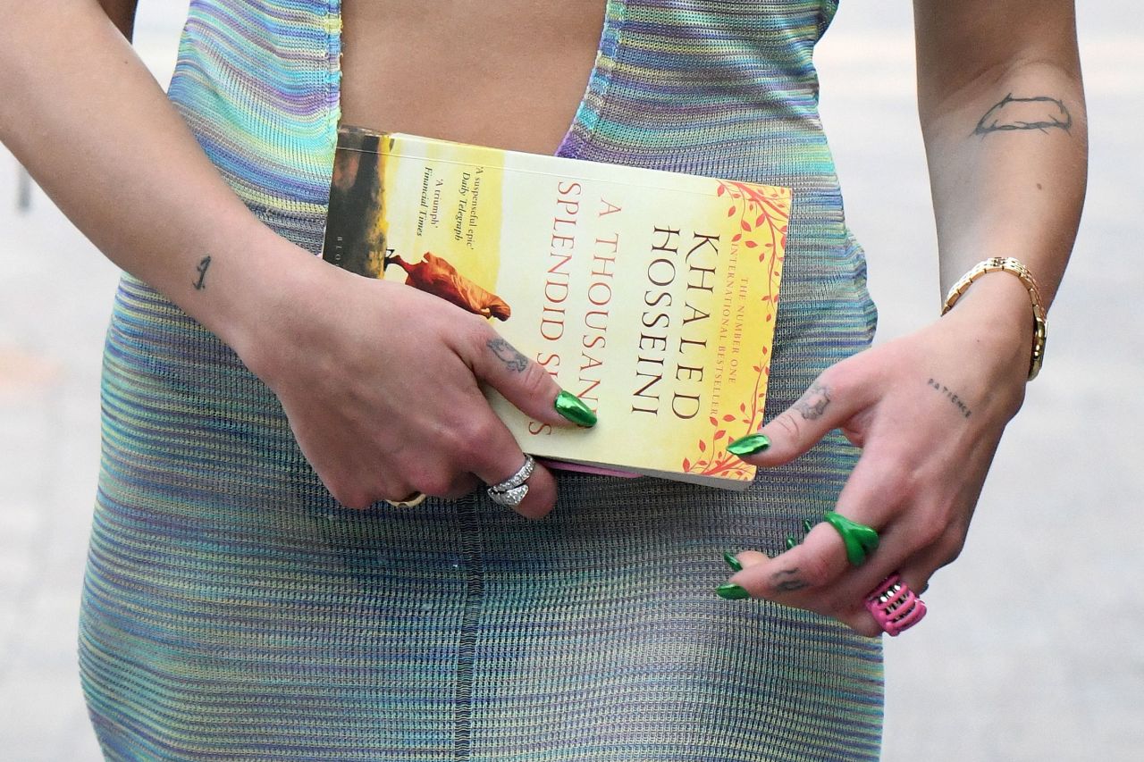 Close-up of tattooed hands holding a copy of 'A Thousand Splendid Suns' by Khaled Hosseini, with green painted nails and rings visible.