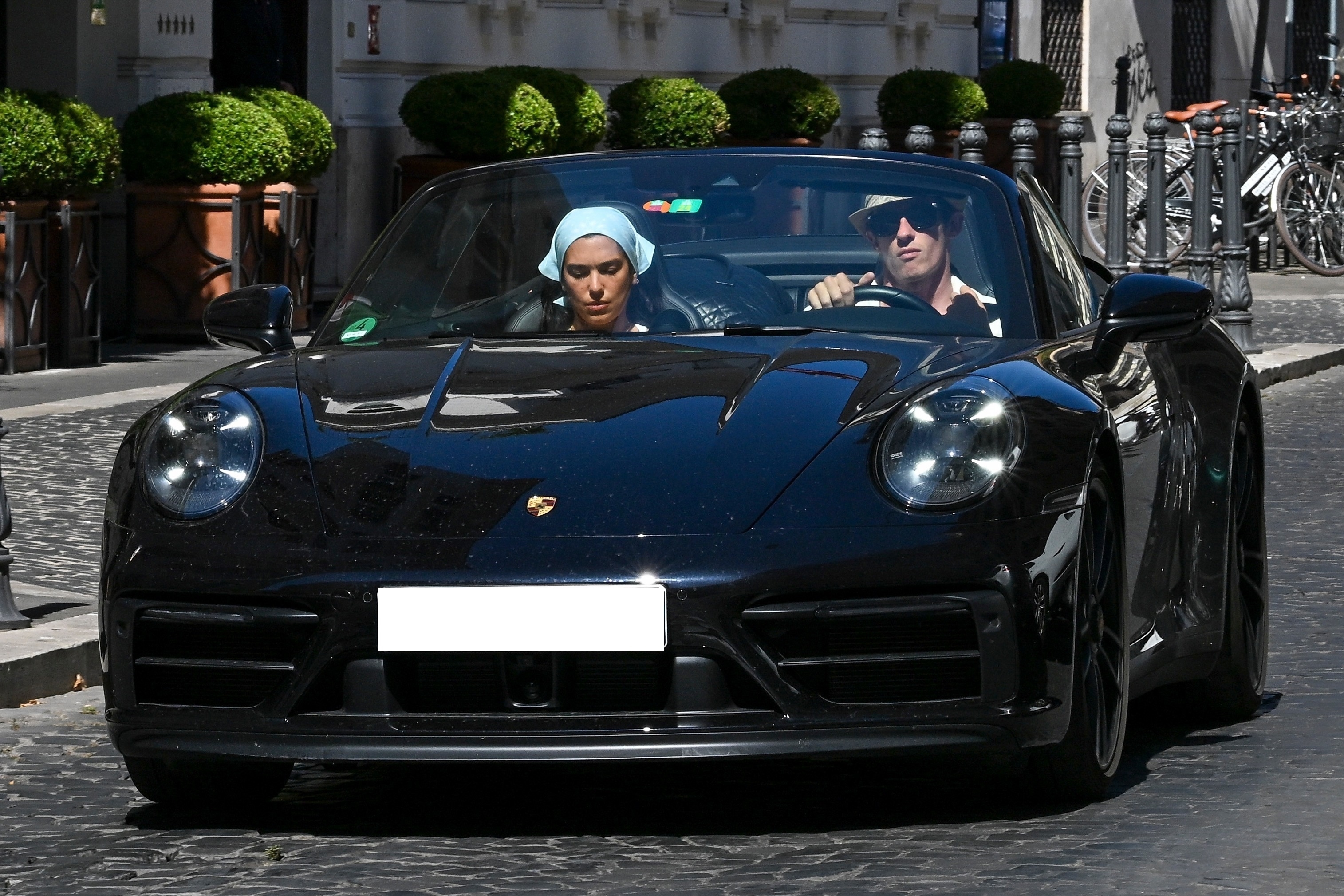 A man in sunglasses and straw hat drives a black Porsche 911 convertible through Rome with a woman in a pale blue headscarf in the passenger seat.