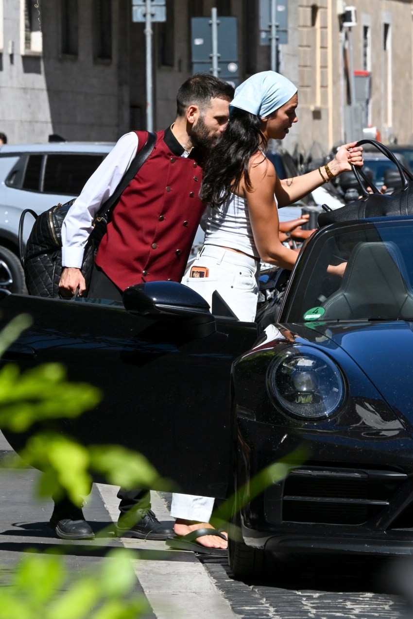 A bearded man in a red vest kisses a dark-haired woman in a white vest top and light blue headscarf next to a black Porsche convertible on a city street.