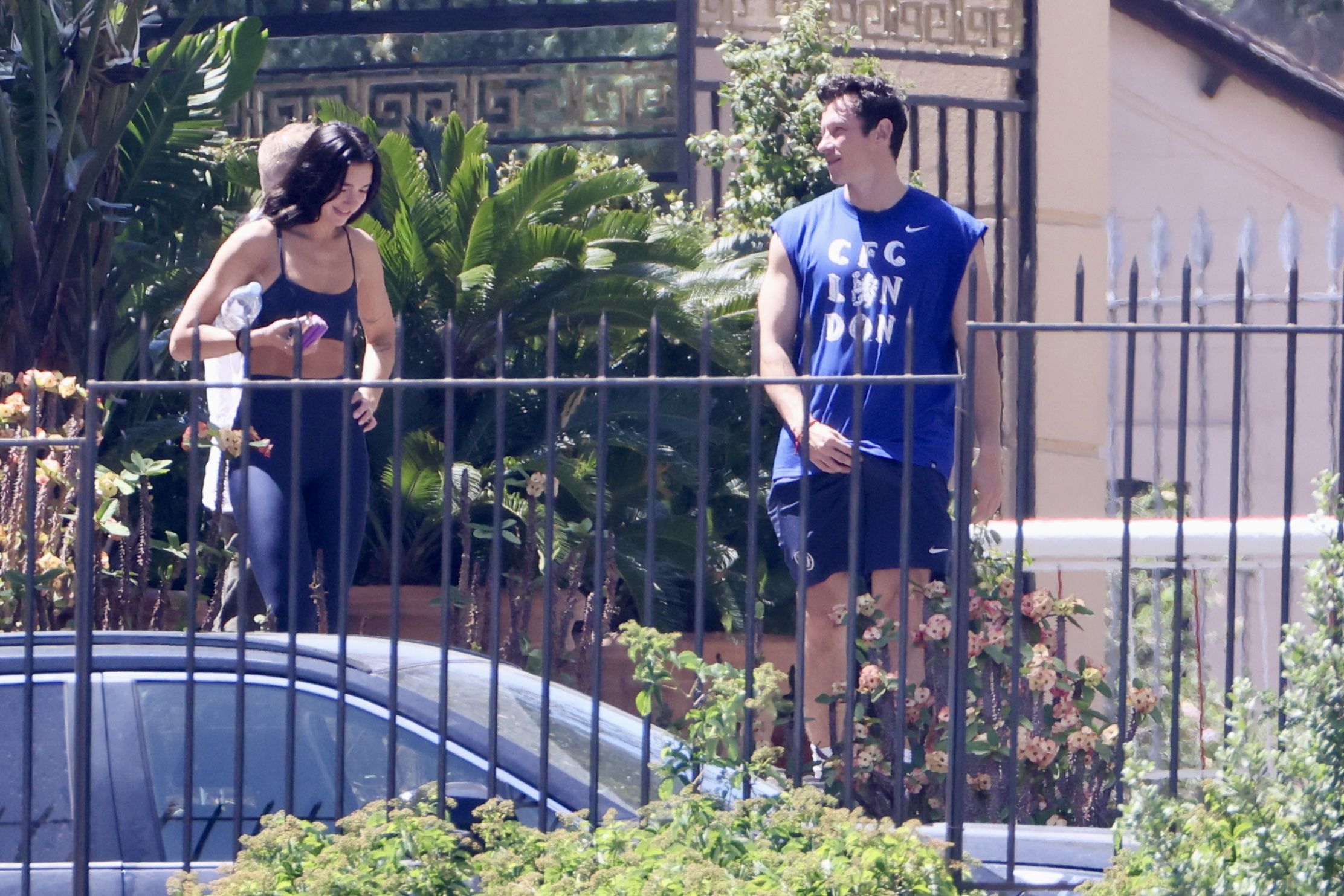A dark-haired woman in navy workout gear and a man in a blue CFC London sleeveless shirt walk together laughing outside a gated property.