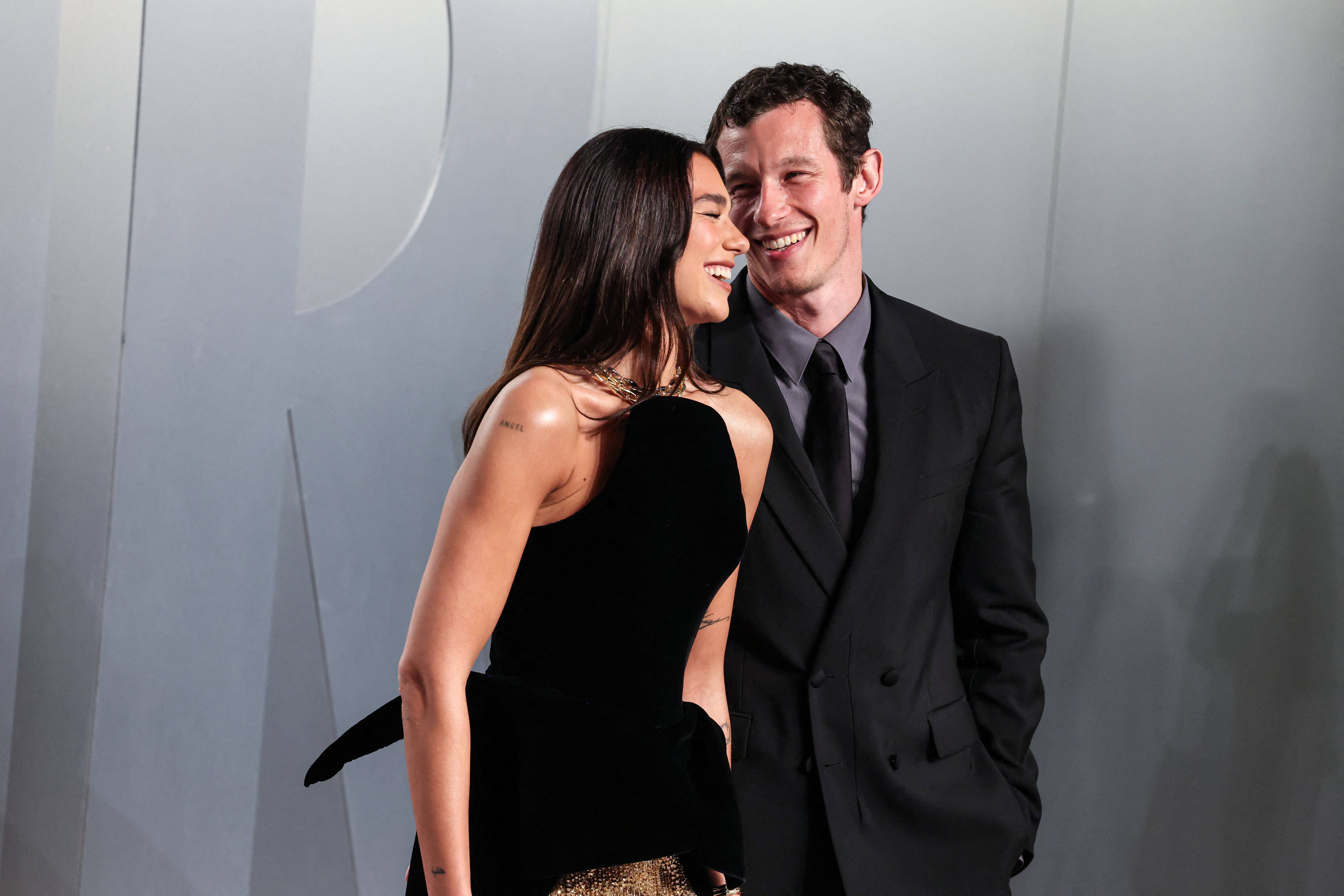 A woman in a strapless black and gold gown laughs while leaning close to a smiling man in a black double-breasted suit against a grey backdrop.