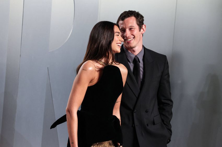 A woman in a strapless black and gold gown laughs while leaning close to a smiling man in a black double-breasted suit against a grey backdrop.