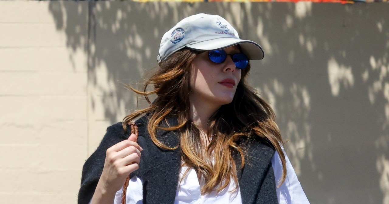 Pregnant Elizabeth Olsen walking outdoors in LA, wearing a white button-down shirt open at the bottom, white trousers, a baseball cap, and blue mirrored sunglasses, carrying a brown woven bag.