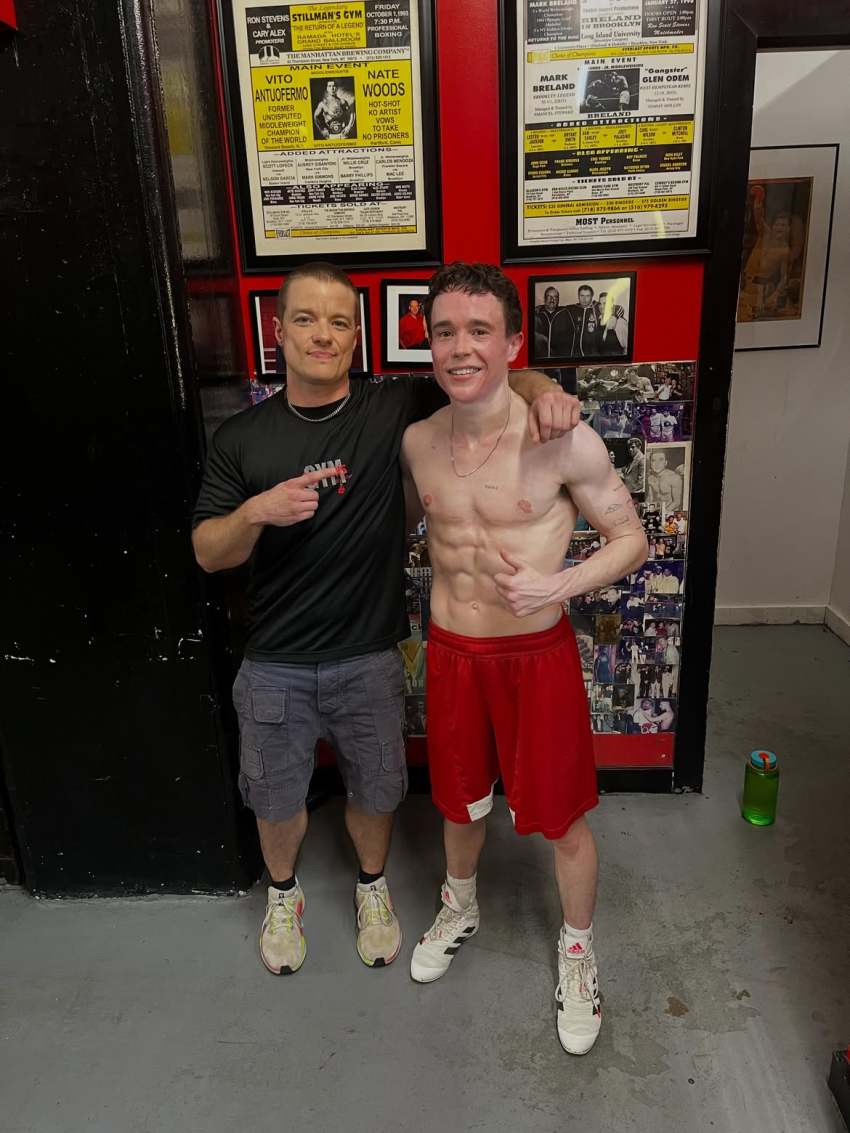Shirtless lean young man in red shorts stands arm-in-arm with a man in a black gym shirt in front of boxing posters and photo collages on a red wall.