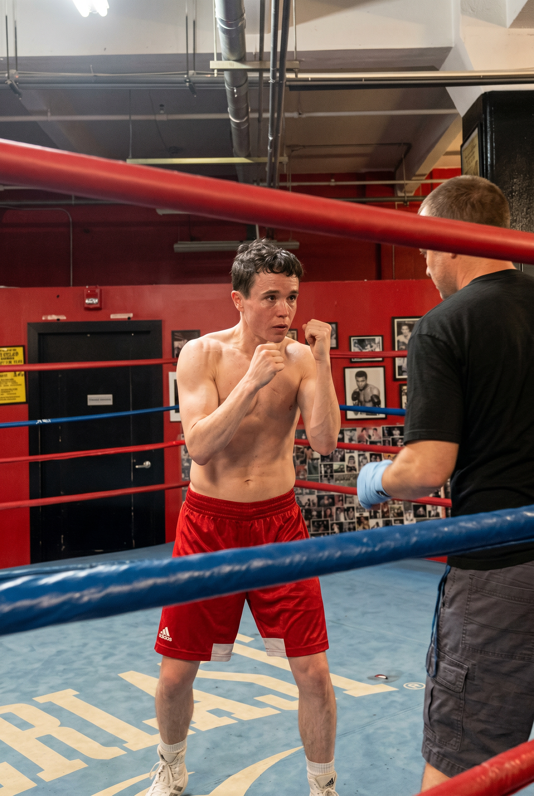 Click to open the image gallery Shirtless young man in red Adidas shorts in a boxing stance with fists raised inside a ring, with a trainer in a black shirt visible at right.