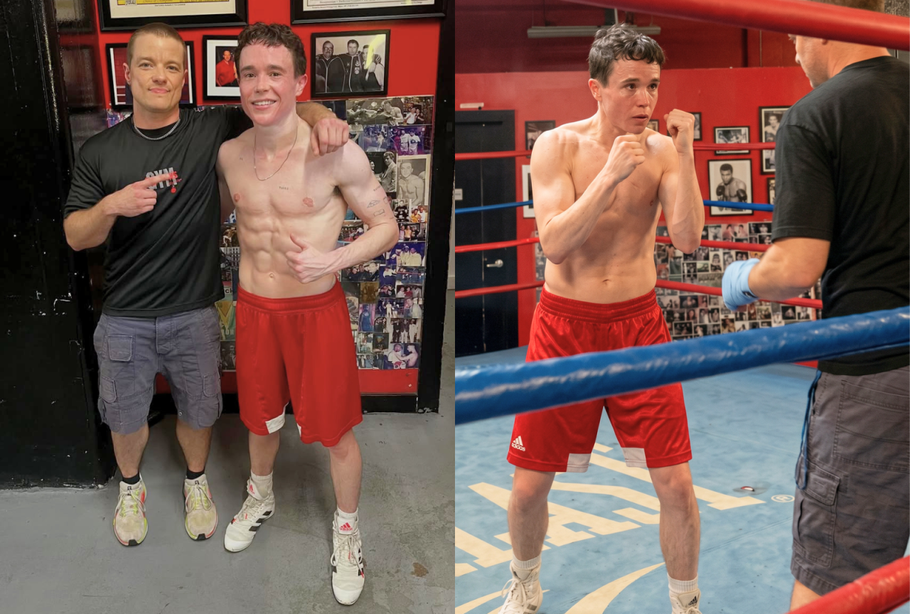 Two men posing together in a boxing gym; one is shirtless in red shorts, the other wears a black gym shirt, with boxing posters on the wall behind them.