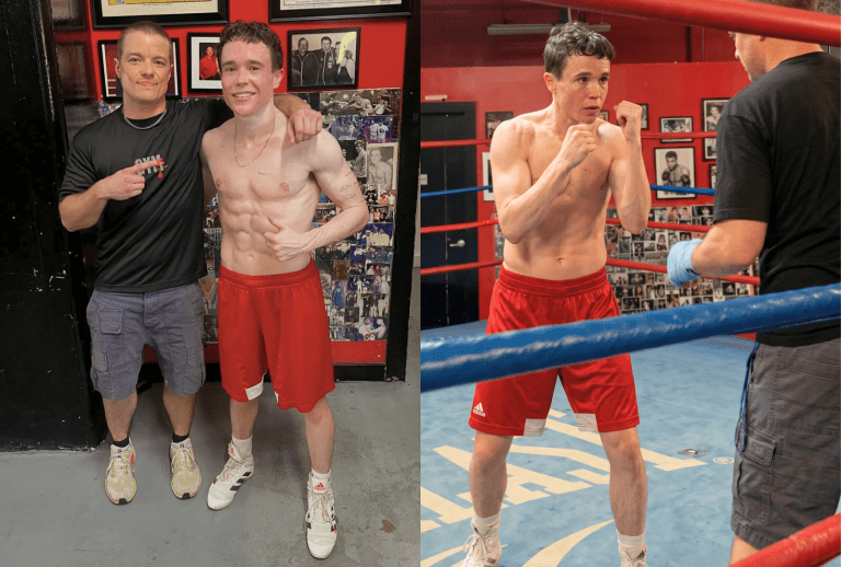 Two men posing together in a boxing gym; one is shirtless in red shorts, the other wears a black gym shirt, with boxing posters on the wall behind them.