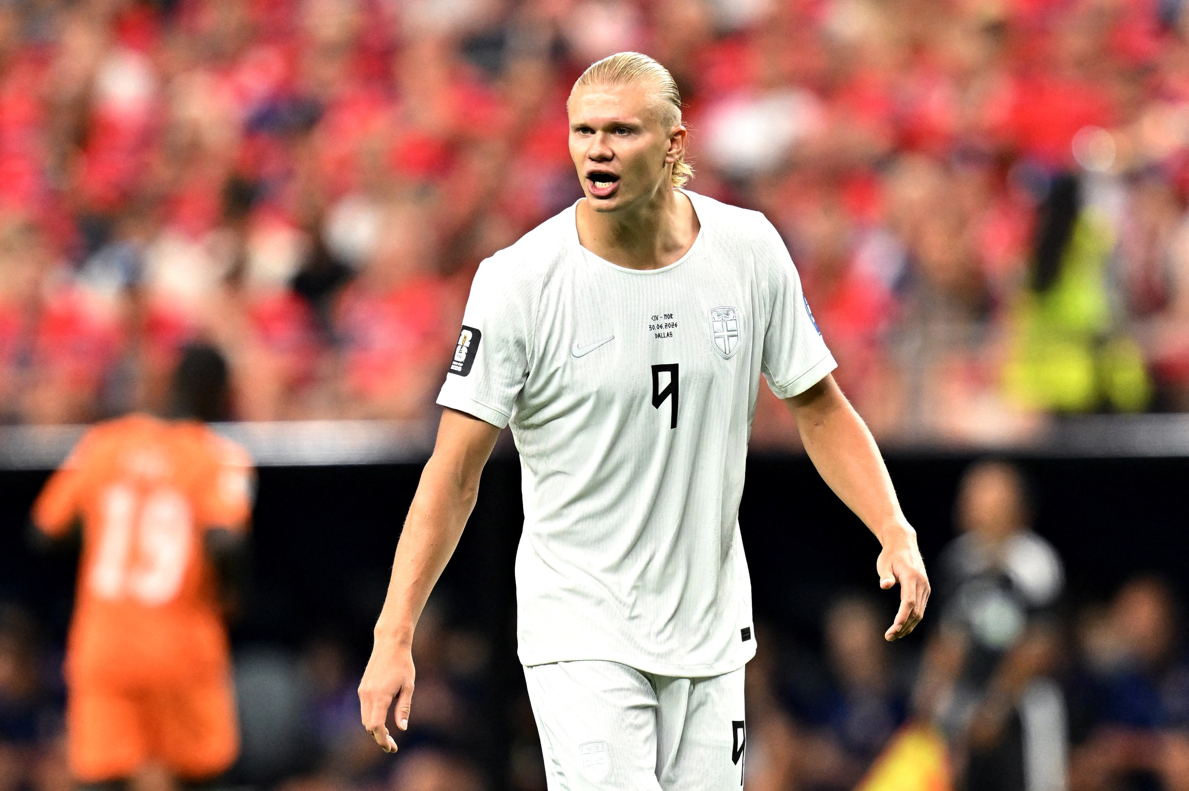 Blond footballer in a white Norway No. 9 jersey walks on the pitch during a World Cup match, crowd in red visible in the background.