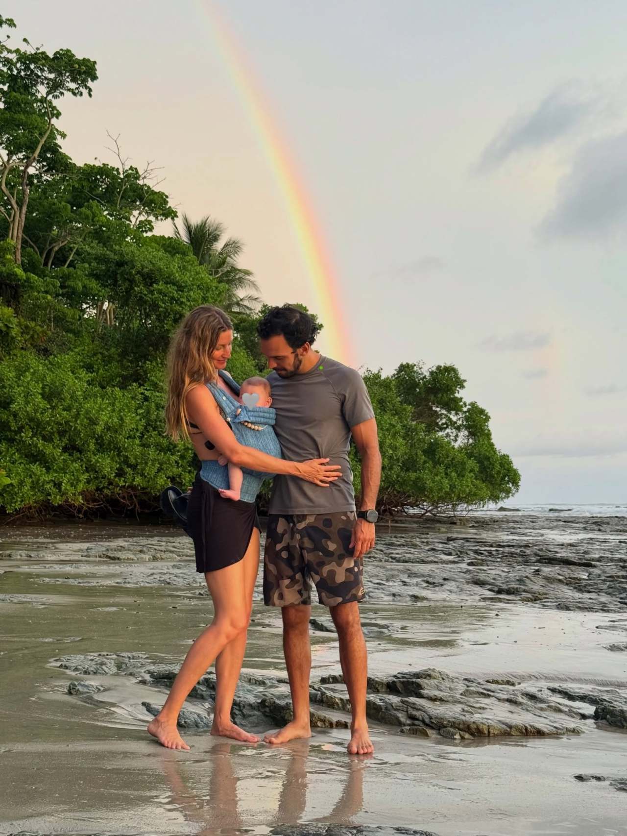 A woman in a black swimsuit holding a baby in a carrier and a man in camo shorts stand together on a rocky beach with a rainbow in the sky behind them.
